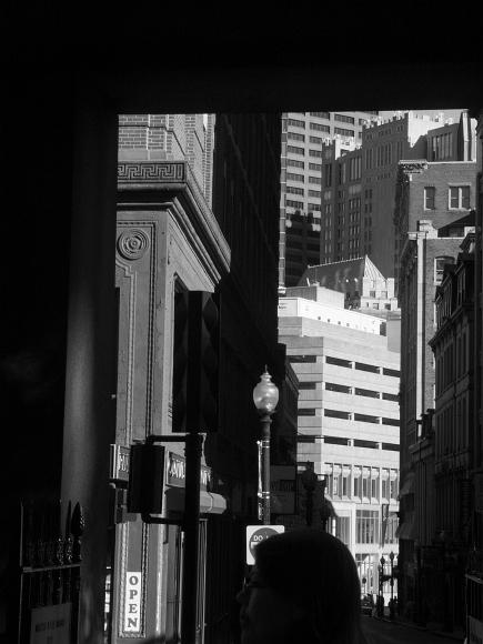Laura peeks into downtown Boston Black and white photograph of a womans head in the foreground looking to the left while layers of tall office buildings, some sunlit and some in shadow lie in...