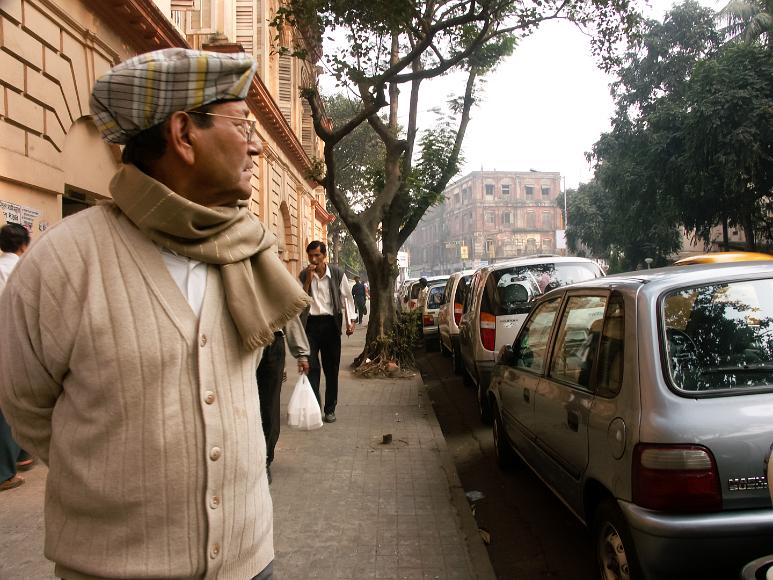 My uncle with a scarf in downtown Kolkata My uncle wearing a sweather, scarf, and checkered hat, standing on the sidewalk in downtown Calcutta, India, looking off to the side past a row of cars