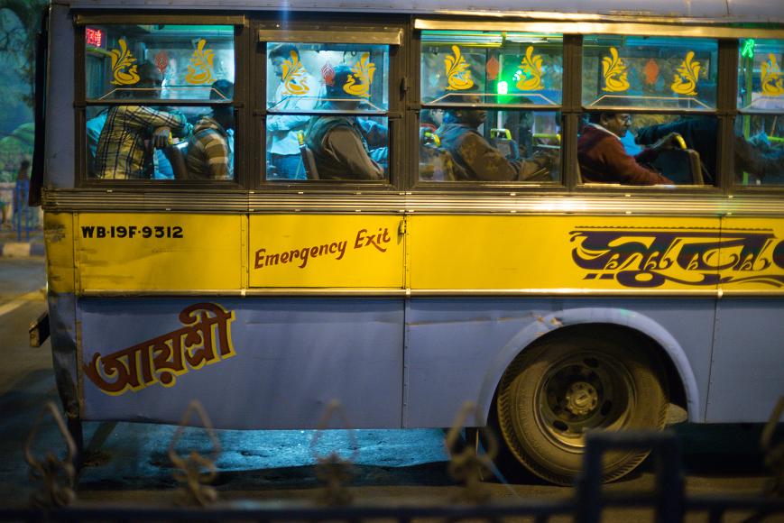 The back of the bus Men sit in the back on a yellow and blue bus at night while a ticket collector goes about his work lit by streetlights
