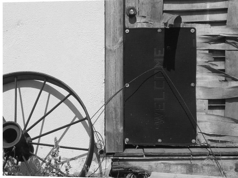Front door of the old Hotel in Nicodemus Kansas Black and white photograph by Rana Banerjee of an old metal tractor or wagon wheel rests against a white stucco wall punctuated by a old wooden door with thin...