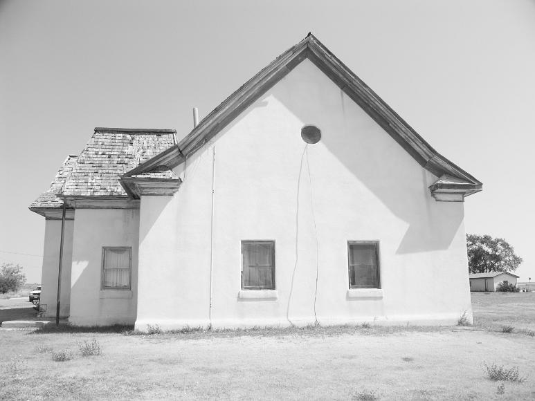 White House in Nicodemus Kansas Black and white photograph of a white stucco building with clapboard shingles on the roof with a car barely visible in the background and done almost entirely...