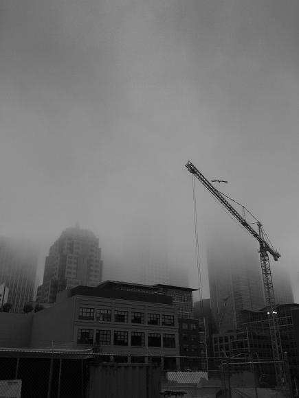 Downtown swallowed by fog Fog covers the tops of buildings while a crane stands silhouetted against the gray sky and fence bounds a construction site below