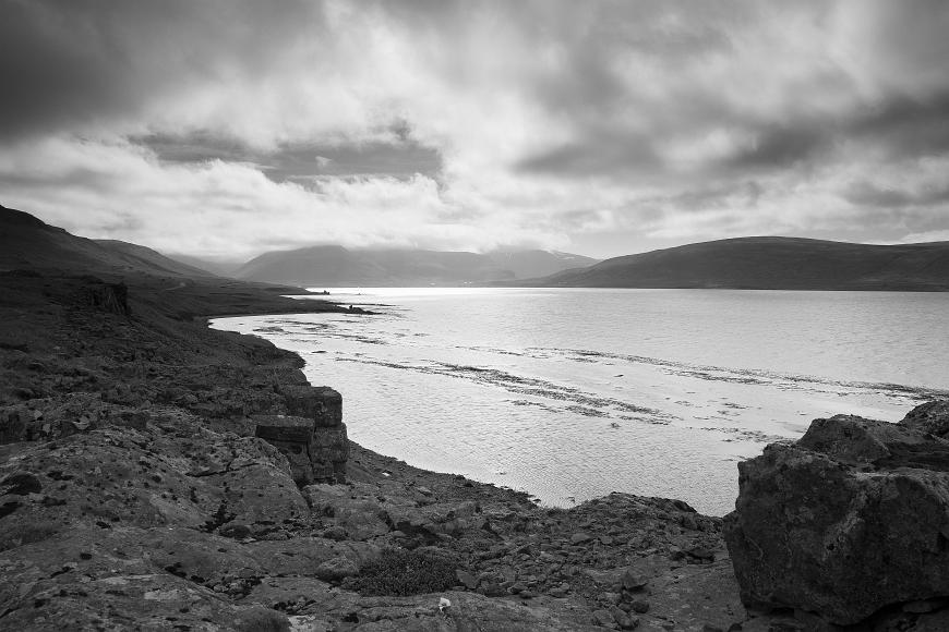 Fjord in Northwestern Iceland Black and white image of a bay and mountains under a partially cloudy sky with a rocky shore dominating the foreground