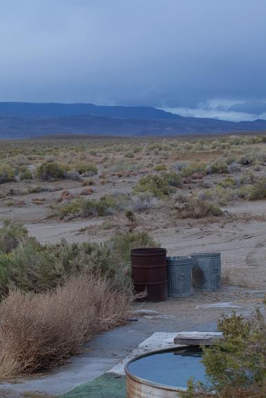 Spence Hot Spring A cattle stock tank is filled with hot water surround by a platform on the desert floor and garbage cans nearby and a cloudy rainy sky set against dark blue...
