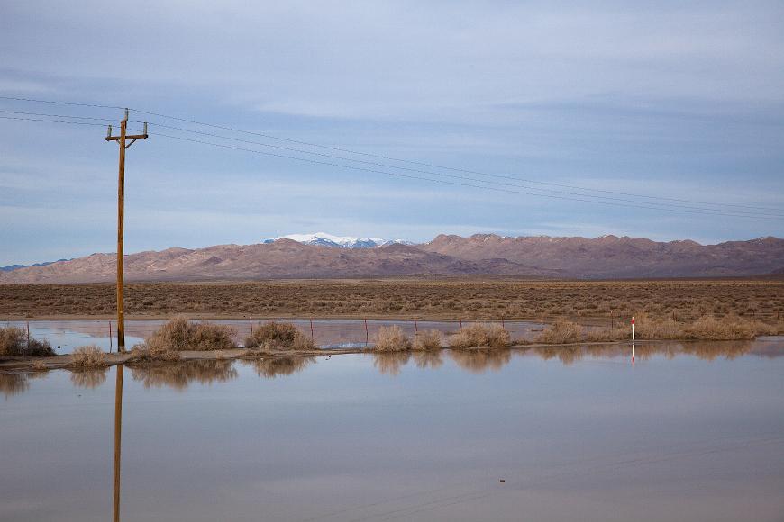 Shallow lake mountains and power lines A shallow lake in a wide valley filled with desert scrub reflects power lines and metal barbed wire fence with a range of barren mountains purple and brown in...