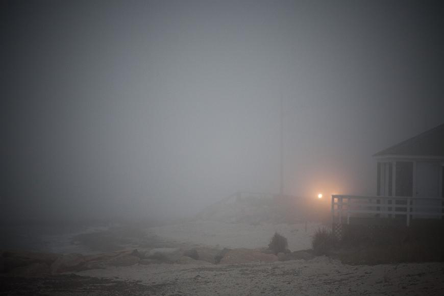 Lightbeam in the fog along the waterfront highway in Woods Hole Cape Cod Massachusetts one yellowed headlight from a car can be seen with the other one hidden by the beck deck of a waterfront house with the beach leading to the ocean which is...