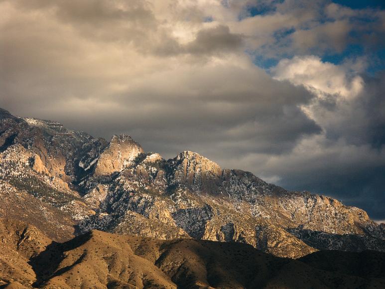 Telephoto shot of Sandia Mountain crest dusted with white and dappled with sun and clouds Telephoto shot of Sandia Mountain crest with granite, limestone and feldspar crags, dusted with white, dappled with sun and clouds and a few small pinyon and...