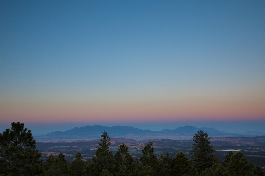 Dusk over trees and sandstone in southern utah dusk sky with the shadow of the moon visible above mountains ranges with varicolored sandstone formations in the midground, and green confiers in the...