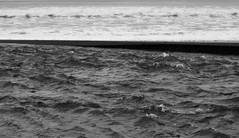 Three layers two water one sand The Mattole river is seen before a sandbank and the pacific ocean waves crashing in the background in black and white