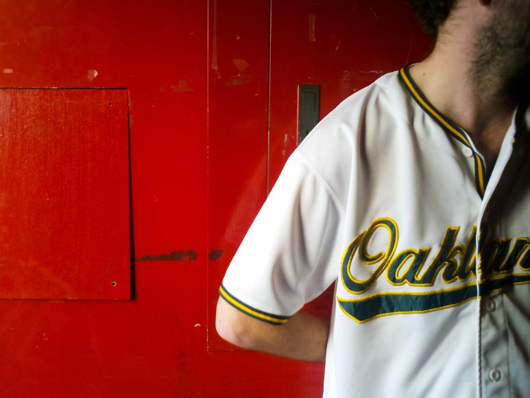Chrispy before the A's game A man wth stubble and his face outside the frame wears an Oakland A's baseball jersey against a bright red wall