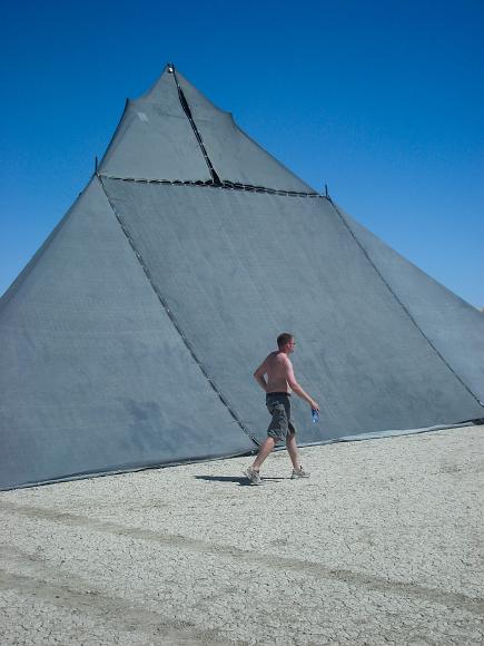 Matt walks in front of Shady at Burning Man 2006 A man walks on a dry lake bed with vehicle tracks in front of a mesh black pyramid in the desert sunlight