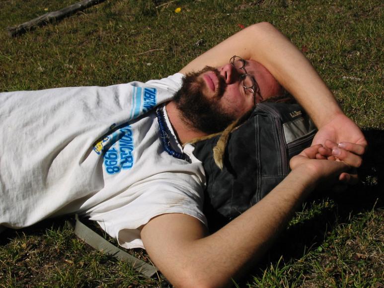 Chris at rest A man with long blond dreadlocks, a glass bead necklace, thin wire-frame glasses, and a tibetan freedom concert 1998 T-shirt, sleeps on grass, using an LL Bean...